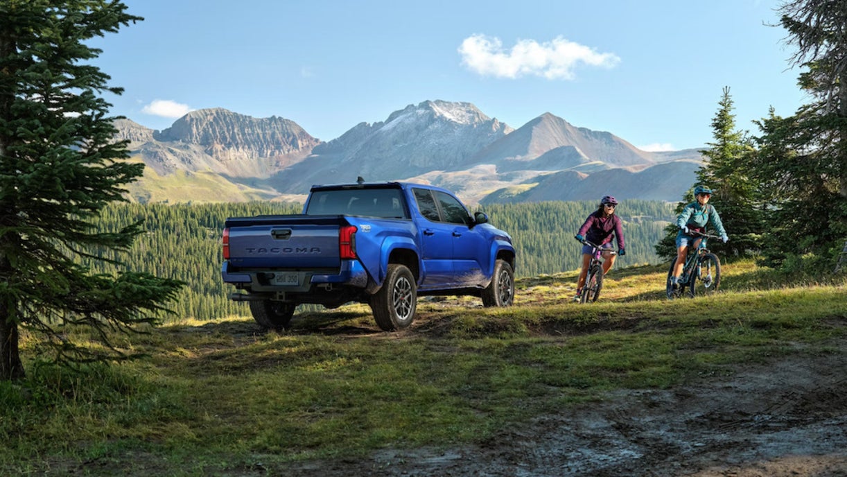 A bright blue 2025 Toyota Tacoma truck parked by a muddy road, with two cyclists passing by and mountains in the distance