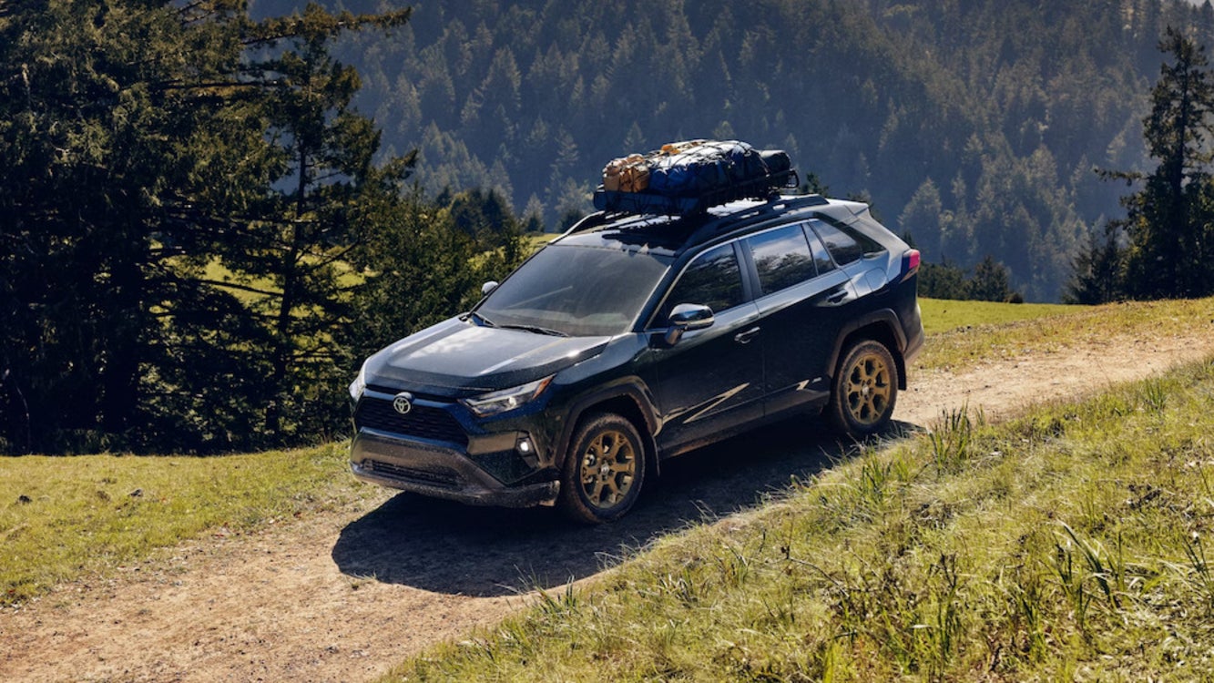 A dark blue 2025 Toyota RAV4 SUV with a packed roof rack driving along a dirt path, with pine trees in the background