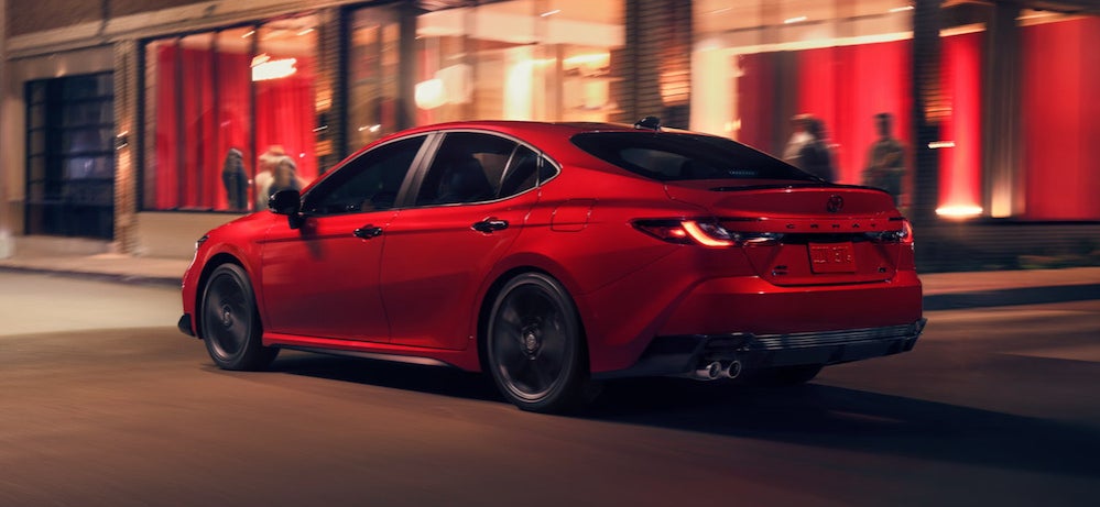 A red 2026 Toyota Camry sedan driving past illuminated storefronts at night