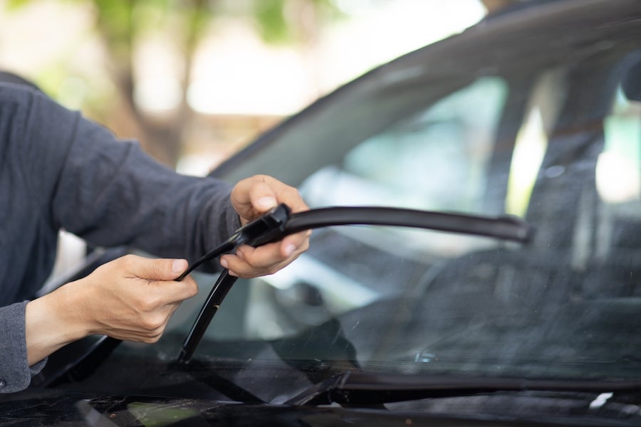 The hands and arms of a person wearing coveralls replacing the windshield wiper blades of a vehicle