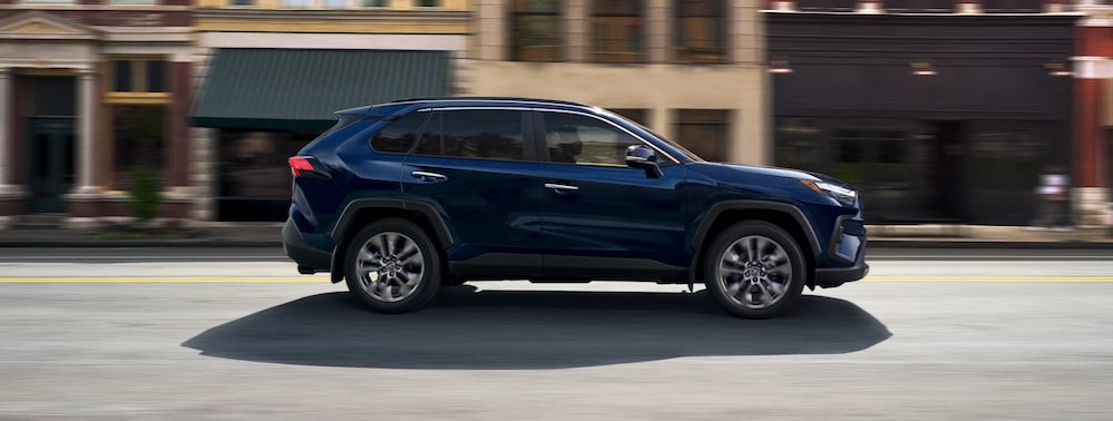 A dark blue 2025 Toyota RAV4 SUV driving down a street past brick buildings