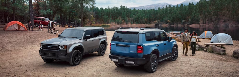 Two 2026 Toyota Land Cruisers, one silver and one blue, parked at a campsite near a lake, with tents and hikers in the background